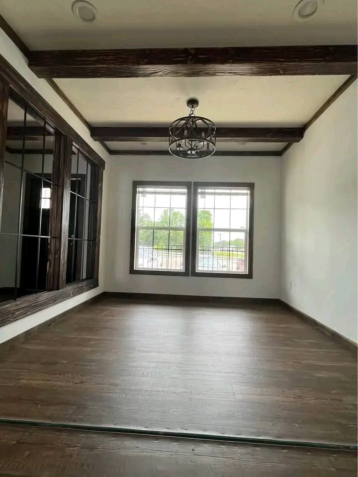 A minimalist room with wooden floors and ceiling beams, featuring large windows and a decorative light fixture. Natural light enhances a calm, inviting atmosphere.