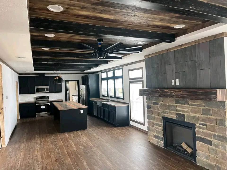Spacious modern kitchen and living area with dark wood accents, sleek black cabinets, large kitchen island, stone fireplace, and ceiling fan.
