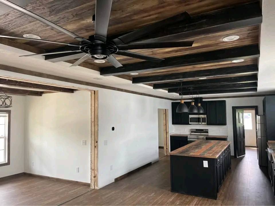 Spacious modern kitchen with dark wood beams, a large ceiling fan, and an island with a wood countertop. Black cabinets and stainless steel appliances add a sleek touch.