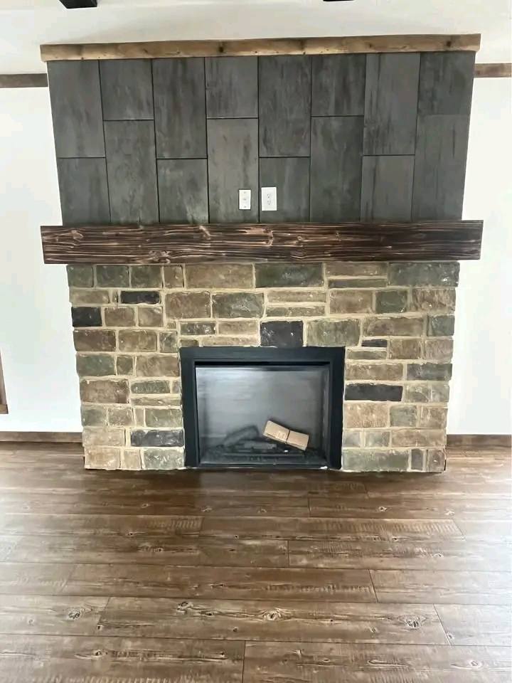 Stone fireplace with a wooden mantel and dark gray textured wall panels. The floor is wooden, creating a rustic and cozy atmosphere.