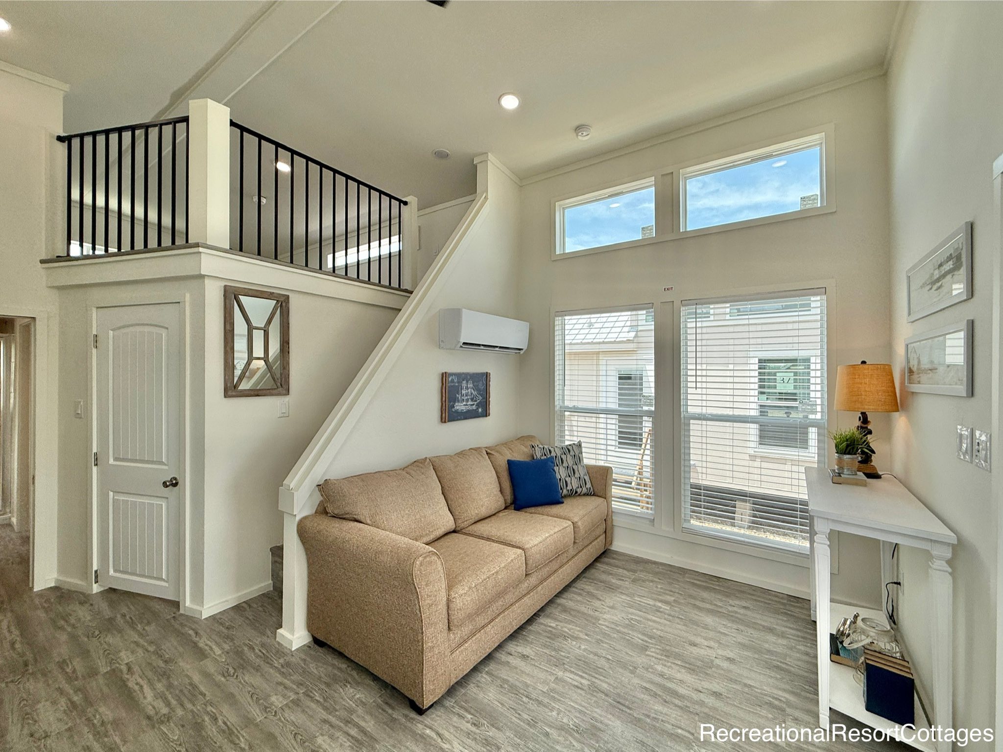 Modern living room with high ceilings and large windows, featuring a beige sofa, a small table with a lamp, and a loft with black railing above.