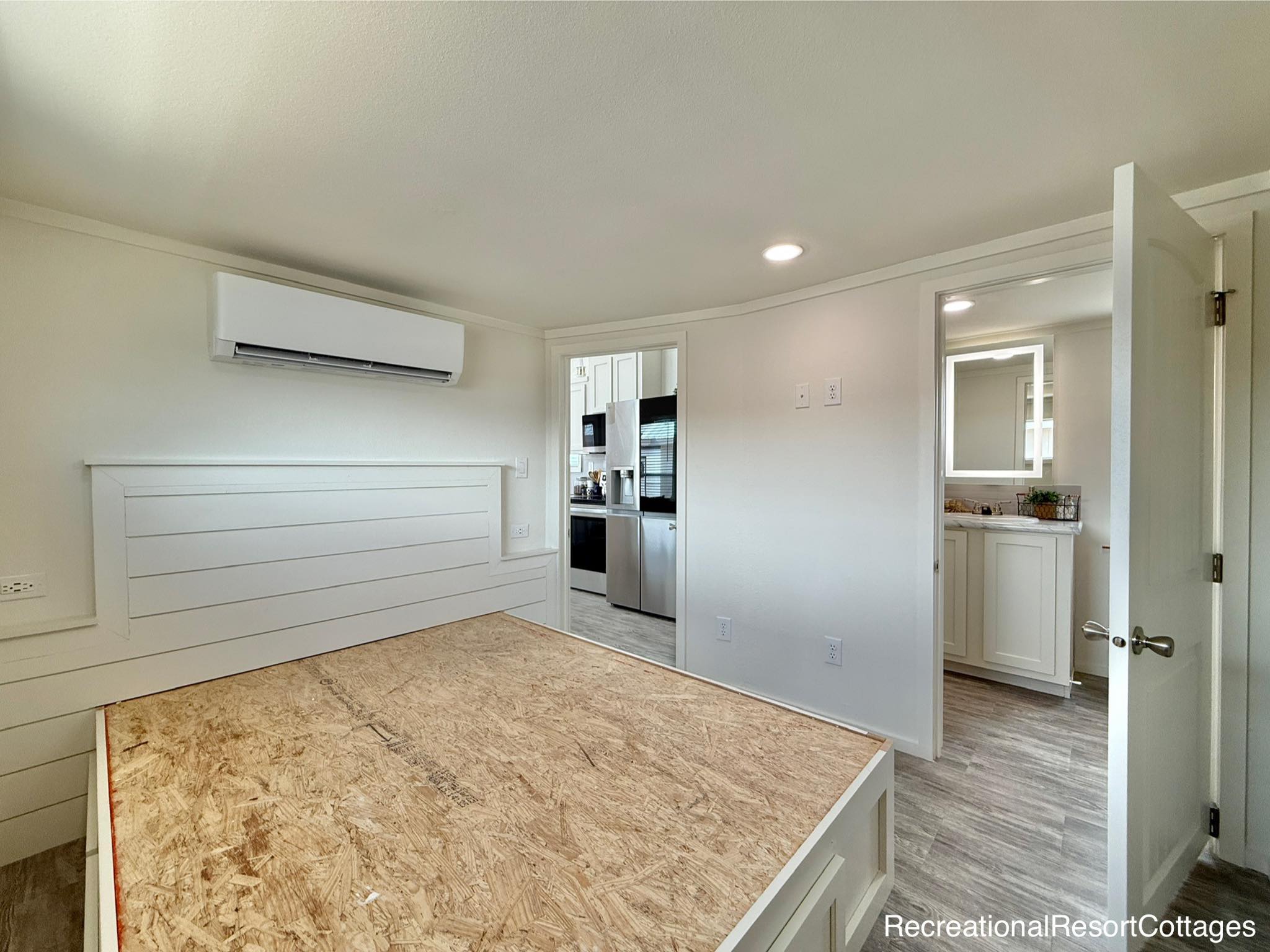Simple white bedroom with unfinished wooden bed frame, shiplap headboard, and wall-mounted air conditioner. Doorway leads to kitchen and bathroom. Bright, minimalistic feel.