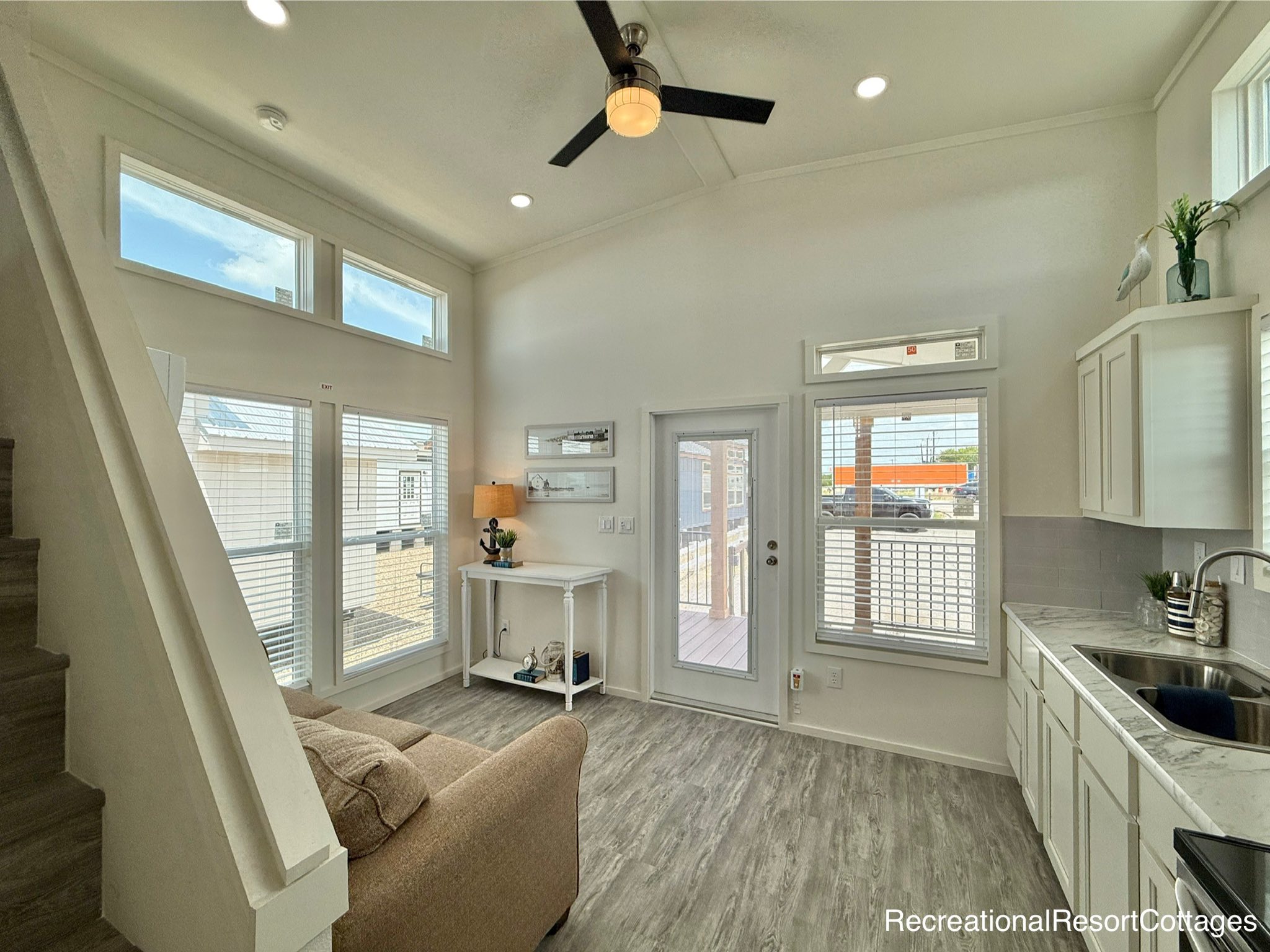 Bright, modern tiny home interior with high ceilings, a ceiling fan, and large windows. Features a cozy sofa, white kitchen cabinets, and wood-look flooring.