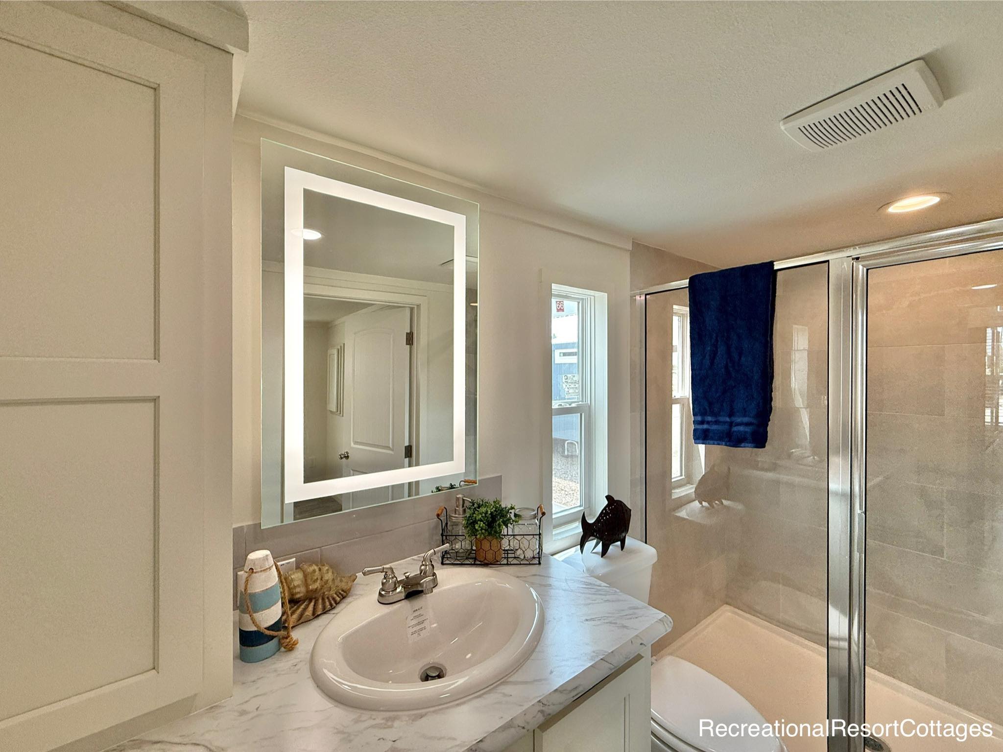 Modern bathroom with a marble countertop and a round sink. Features an illuminated mirror, a glass shower with a blue towel, and minimal decor.