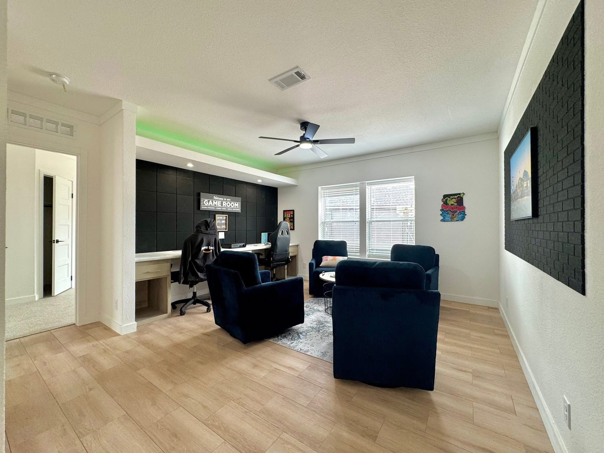 Modern game room with wooden floor, navy armchairs, and a gaming setup under a "Game Room" sign. Bright with natural light from windows.