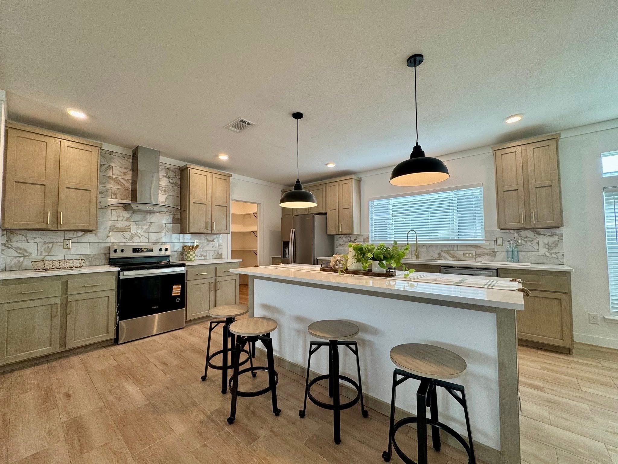 Modern kitchen with wooden cabinets, marble backsplash, and stainless steel appliances. A large island with stools and pendant lights adds elegance.