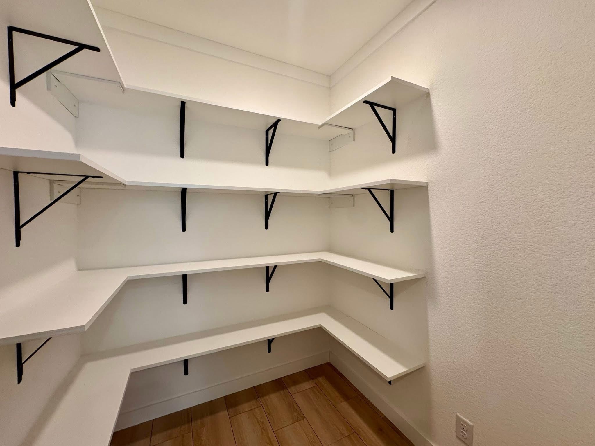Empty pantry with white corner shelves supported by black brackets. The space has a wooden floor and white walls, conveying a clean, minimalist tone.