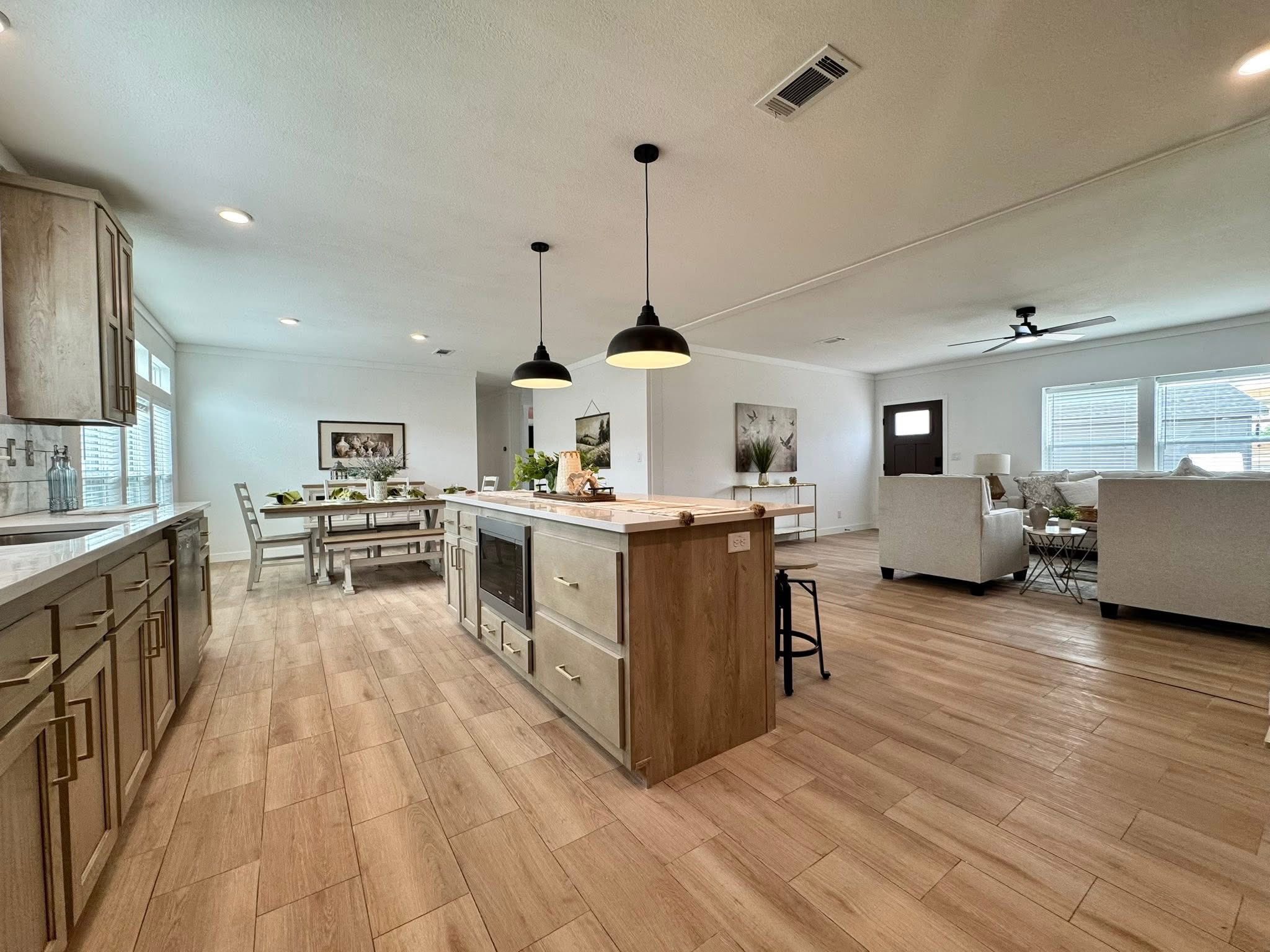 Modern open-concept living space with light wood flooring, featuring a kitchen island with pendant lights, dining table, and a cozy seating area with sofas.
