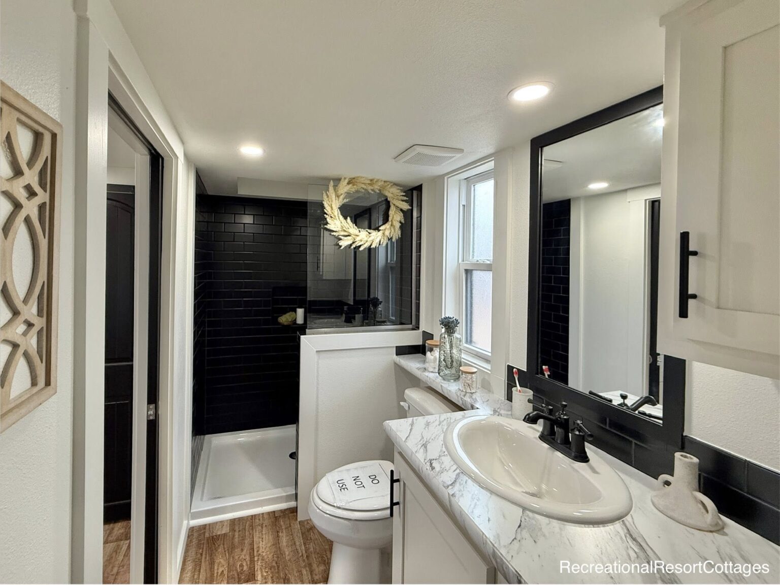 Compact modern bathroom with black-tiled shower, white fixtures, and wood accents. A wreath hangs above, adding a touch of elegance. Warm, inviting tone.