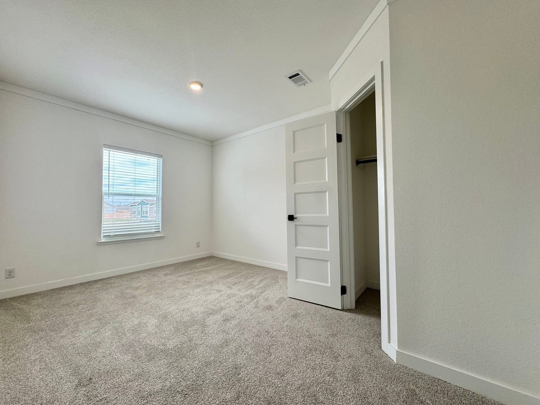 Empty room with beige carpet, white walls, and a window showing a cloudy sky. A small closet with an open door is visible on the right. Minimalistic and clean.