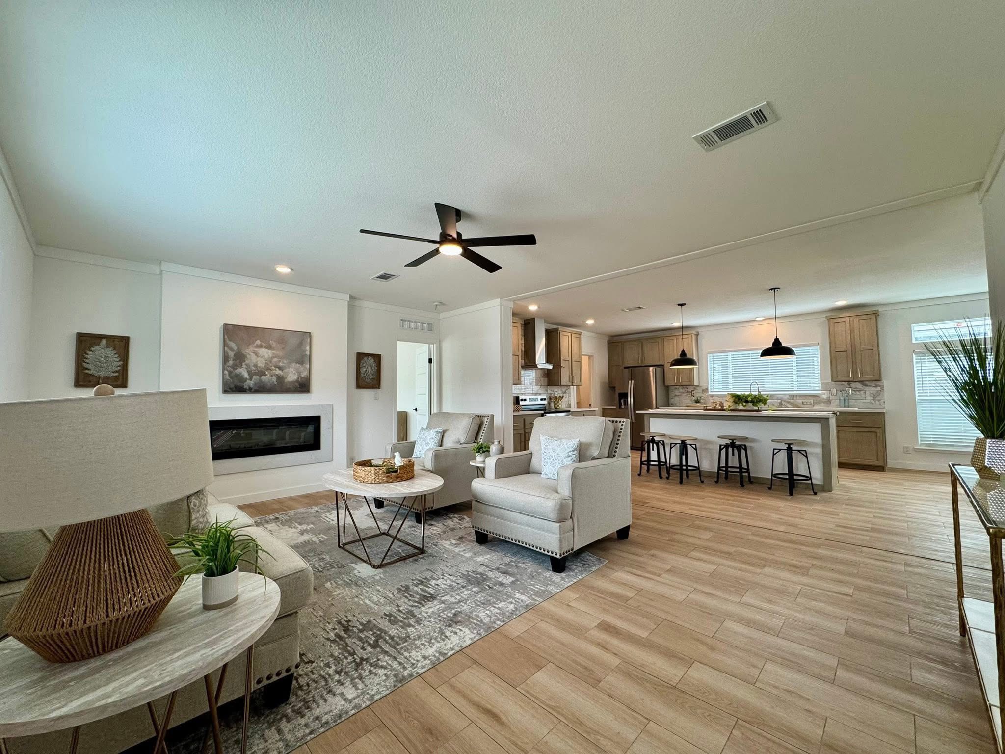 Spacious open-plan living area with light wood flooring, modern furniture, and neutral tones. Kitchen island with bar stools, ceiling fan overhead. Cozy, inviting atmosphere.