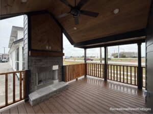 Covered outdoor patio with a wooden ceiling fan, stone fireplace, and wood railings. Overcast sky and road visible in the background. Cozy atmosphere.