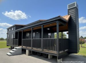 Stylish black tiny house on wheels with a wraparound porch and chimney, set under a bright blue sky. Modern and cozy outdoor vibe.