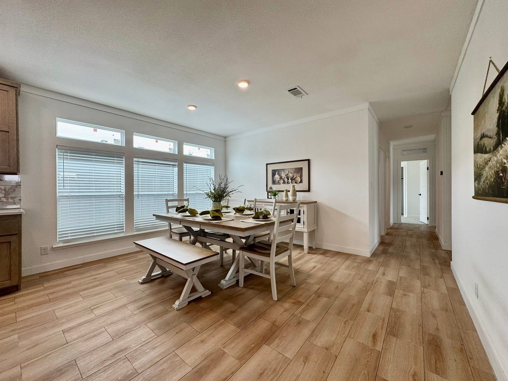 Spacious dining area with a rustic wooden table, white chairs, and a bench. Large windows provide natural light, enhancing the room's warm, inviting atmosphere.