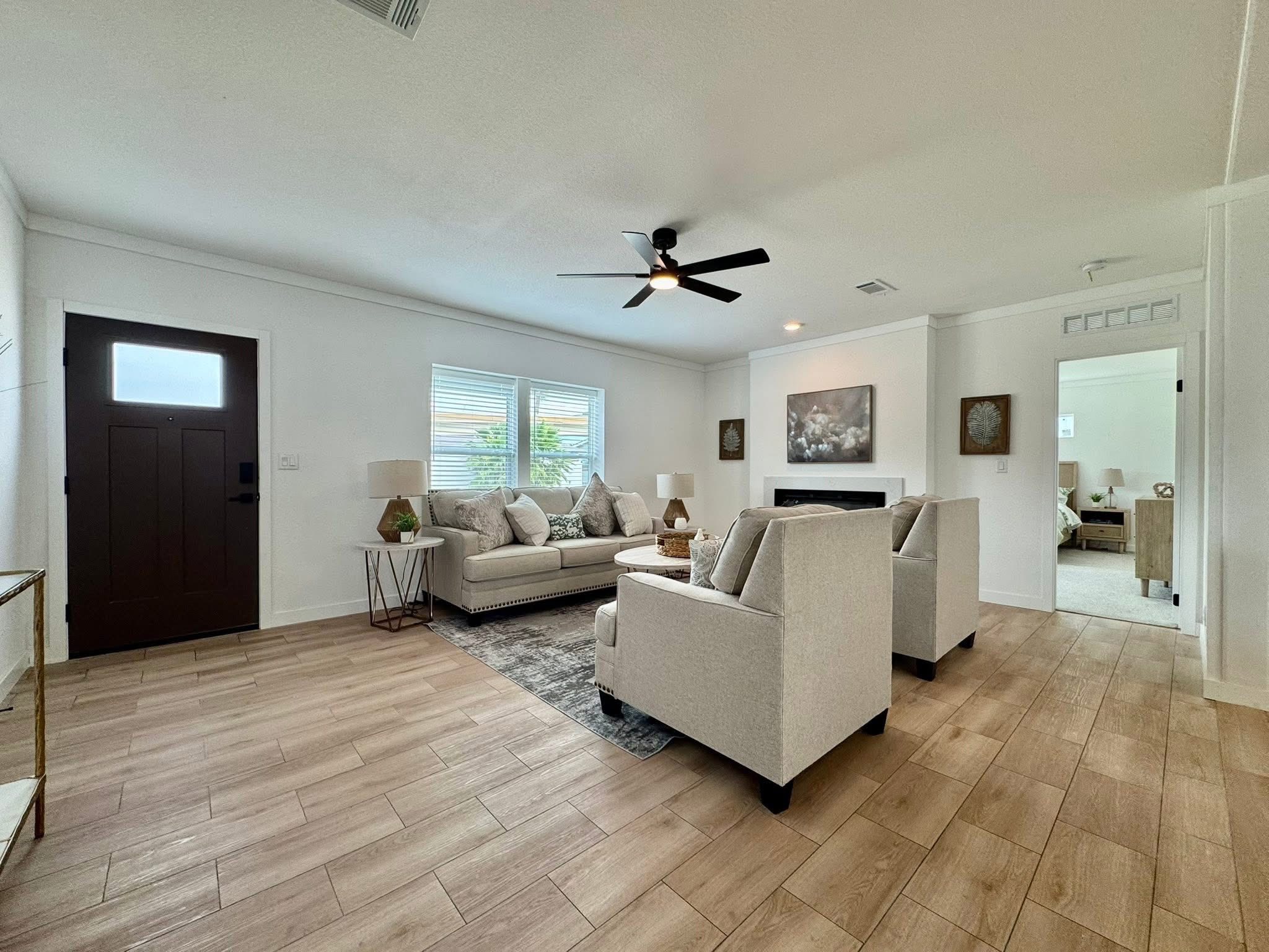 Bright living room with wooden floors, white walls, and a ceiling fan. Grey sofas are arranged around a coffee table, and a large window lets in natural light.