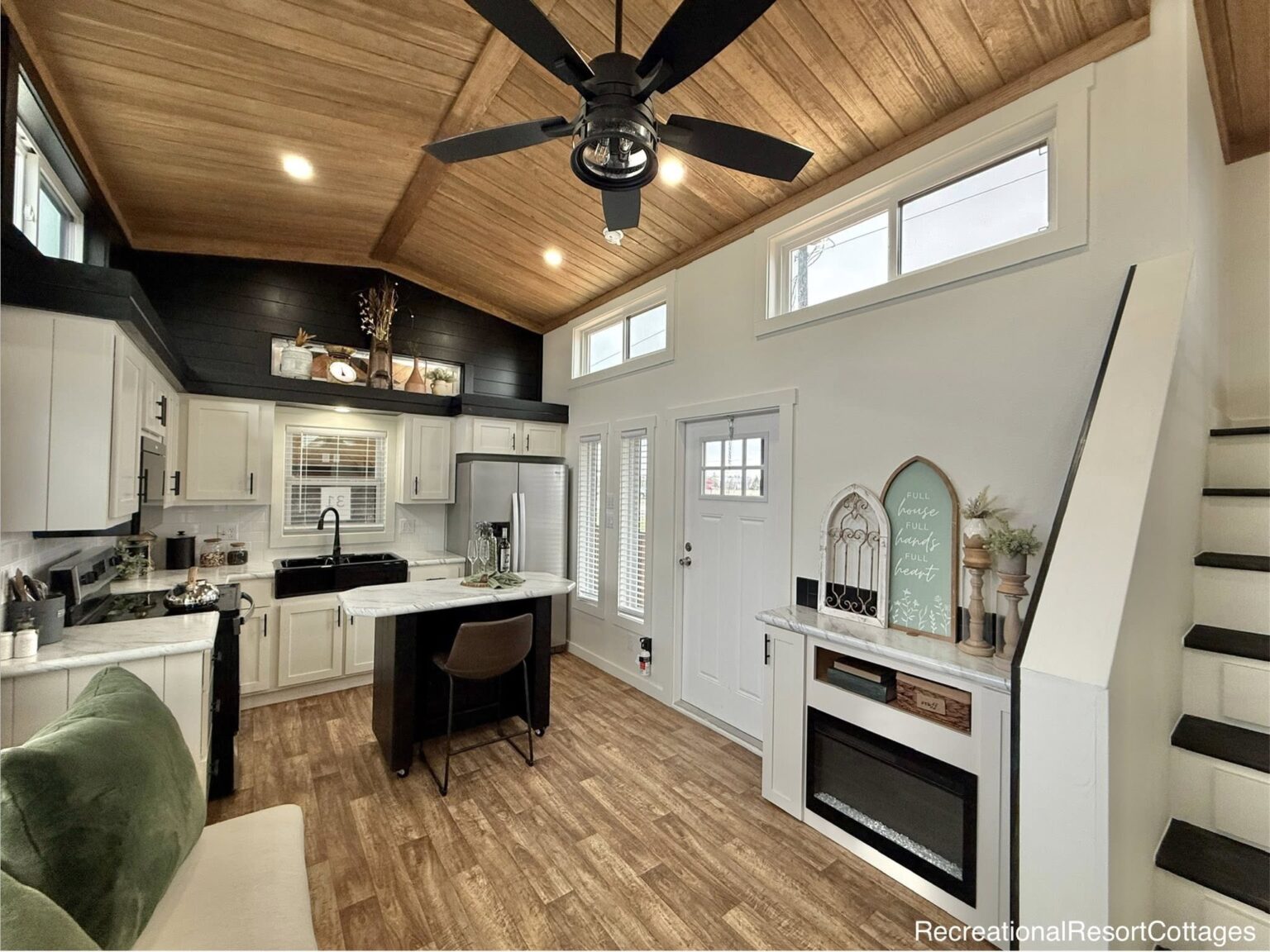 Modern tiny home interior with vaulted wood ceiling, ceiling fan, white kitchen cabinets, island with seating, and cozy decor. Warm, inviting ambiance.