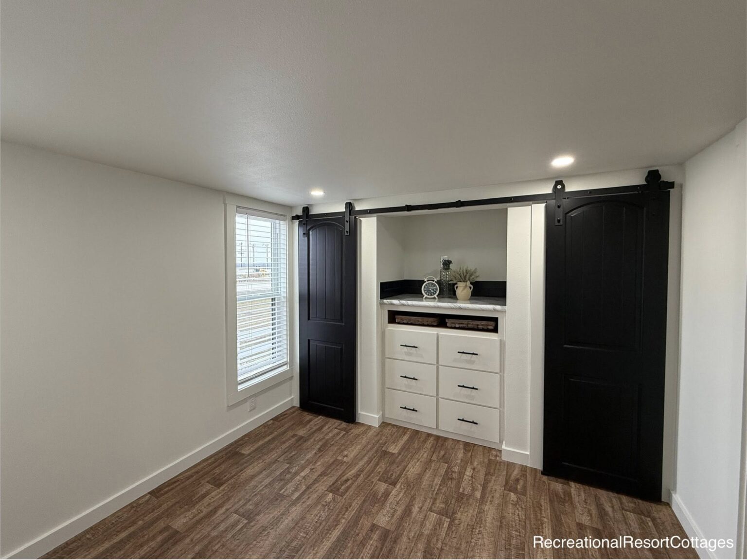 A cozy, modern room with wood flooring features a white built-in dresser under a shelf. Black sliding barn doors and a window with blinds enhance the space.