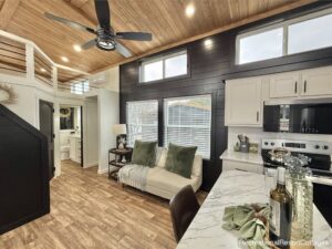 Cozy tiny home interior with wood ceiling, black shiplap walls, and large windows. Modern kitchen, loft space, and living area with green accents.