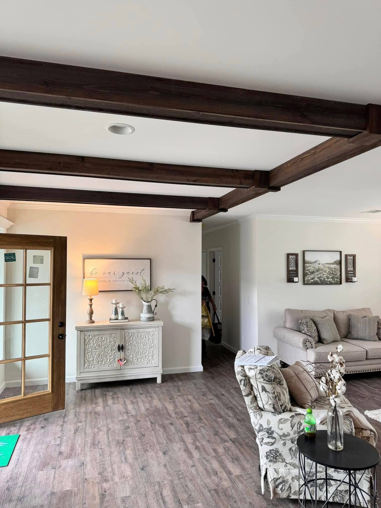 Modern living room with wooden beams and vinyl flooring. Features a beige sofa, floral armchair, and side table with decor. Cozy and inviting atmosphere.