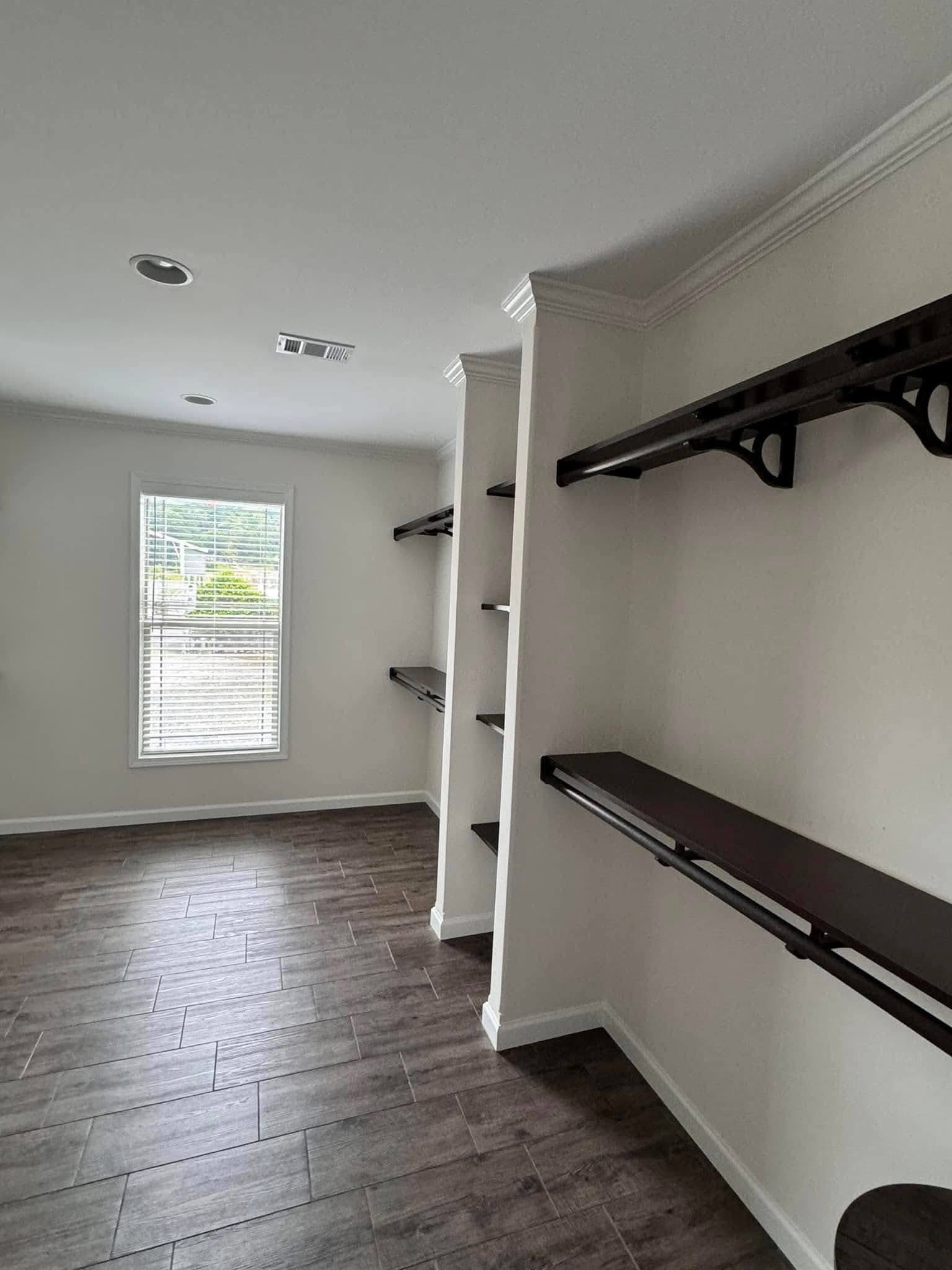 Empty walk-in closet with dark wooden shelves and rods on white walls, wood-patterned floor tiles, and a window letting in natural light.