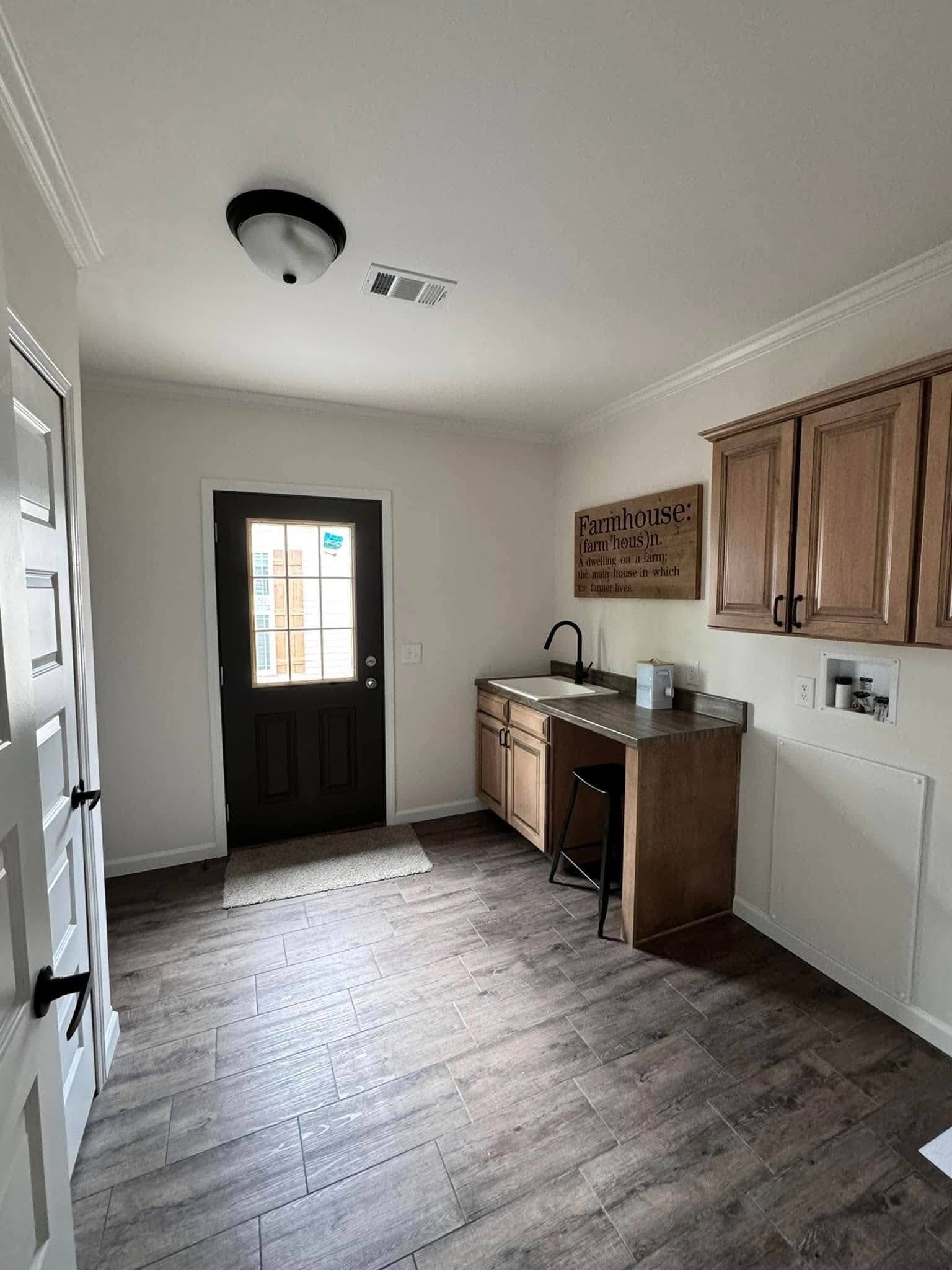 Clean laundry room with wood flooring, beige cabinets, and a farmhouse-style sink. A black back door lets in light, creating a cozy atmosphere.
