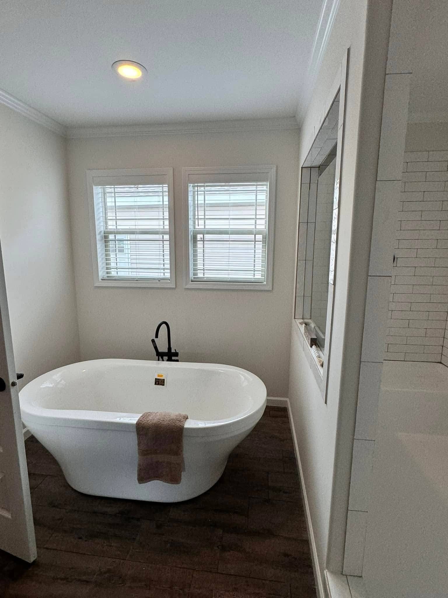 Minimalist bathroom with a white freestanding tub on dark wood floor, black faucet, towel draped over the side, large windows, serene and modern.