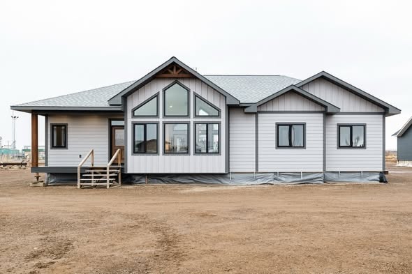 Single-story, modern prefab home with large front windows and a gable roof, set on a barren, dirt-covered plot under a bright, overcast sky.