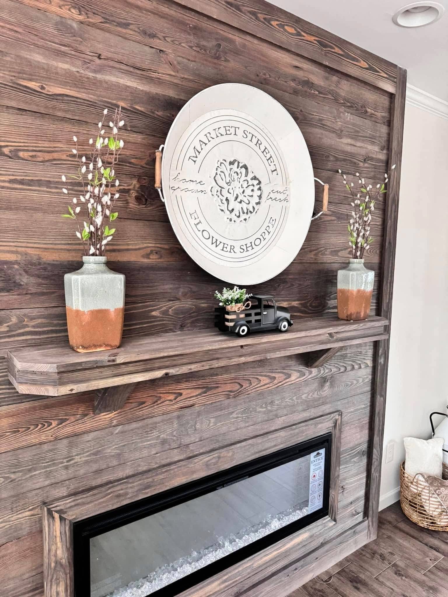 Wall with rustic wooden paneling, featuring a round "Market Street Flower Shoppe" sign. Shelves hold vases with pussy willow branches and a small vintage truck decor. A modern, rectangular electric fireplace is fixed below.