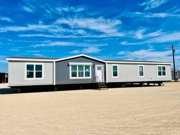 A large, modern mobile home sits on a sandy lot under a bright blue sky with scattered clouds, conveying a sense of openness and tranquility.