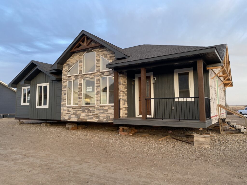 Modern house under construction, featuring dark siding, stone facade, large windows, and a covered porch, set against a cloudy blue sky.