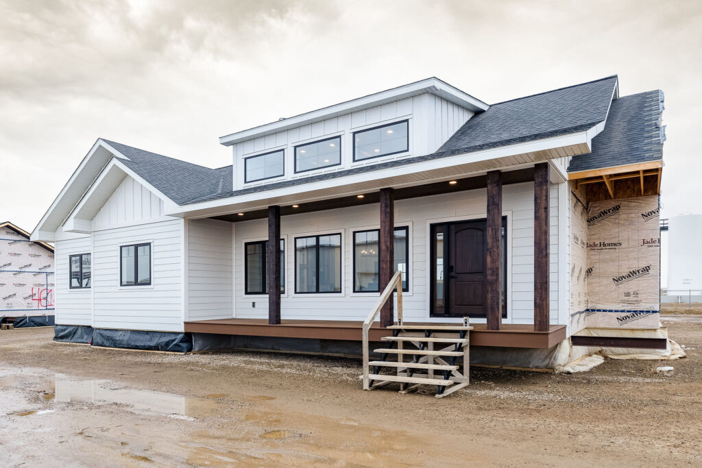 A newly constructed, modern white house with a dark front door, large windows, and a covered porch supported by wooden beams sits on a muddy plot.