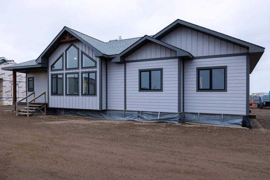 A gray, modern modular home is set against a cloudy sky. Features include large triangular windows and dark trim, with a dirt yard in the foreground.