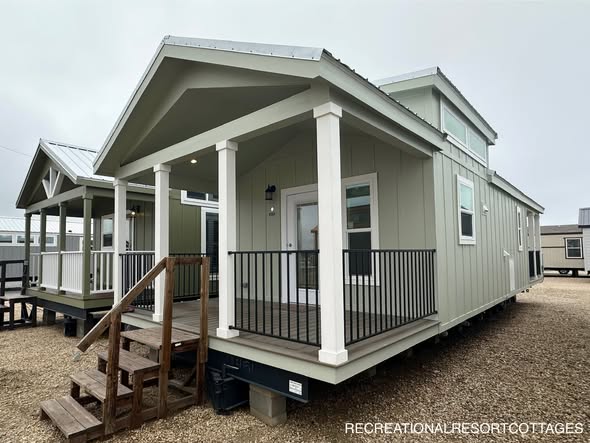Compact green tiny house with a white-trimmed gable roof, elevated wooden porch, and railings. Adjacent similar house visible under a cloudy sky.