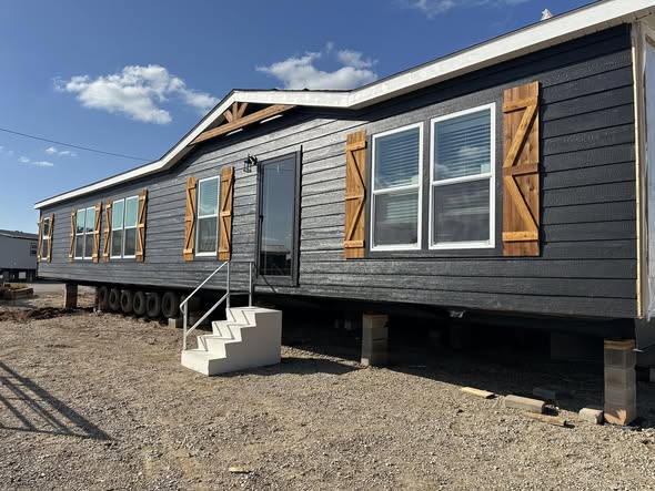 Long dark gray manufactured home with white trim and wooden shutters, elevated on blocks. A small staircase leads to the front door under a sunny sky.