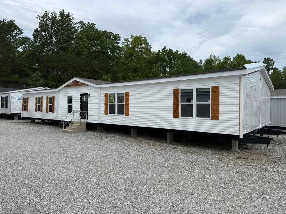 A white mobile home with brown shutters sits on a gravel lot. Surrounded by trees, the scene is calm and the sky is overcast, conveying a serene atmosphere.
