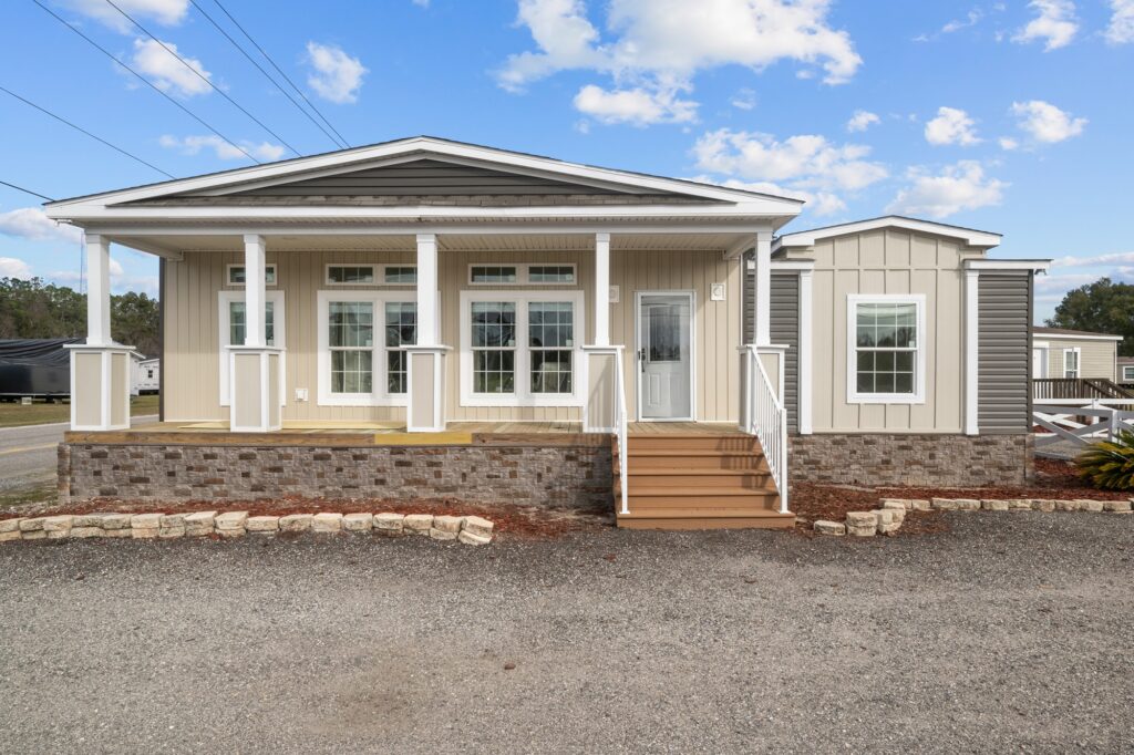 A modern, single-story manufactured home with beige siding and a stone foundation. Features a porch with white columns under a blue sky with clouds.