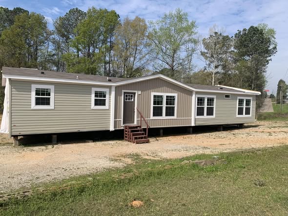 A beige mobile home with white trim is set on a grassy lot, surrounded by trees under a partly cloudy sky. It has multiple windows and a small front porch.