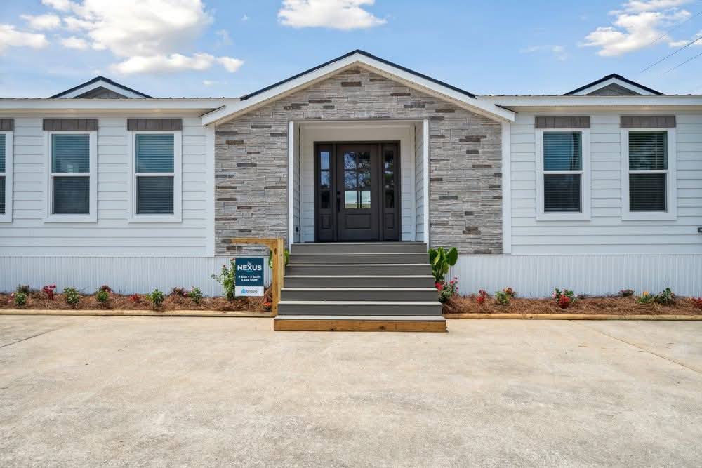 Modern house front with gray stone facade, dark double doors, and six windows. A small garden lines the path, conveying a welcoming atmosphere.