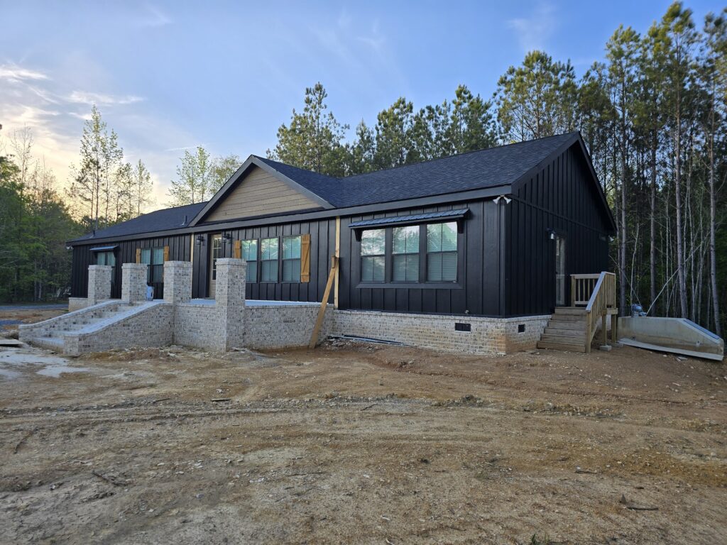 Modern, single-story house with dark siding and large windows, surrounded by trees. Dual steps lead to the entrance, evoking a serene, rustic vibe.