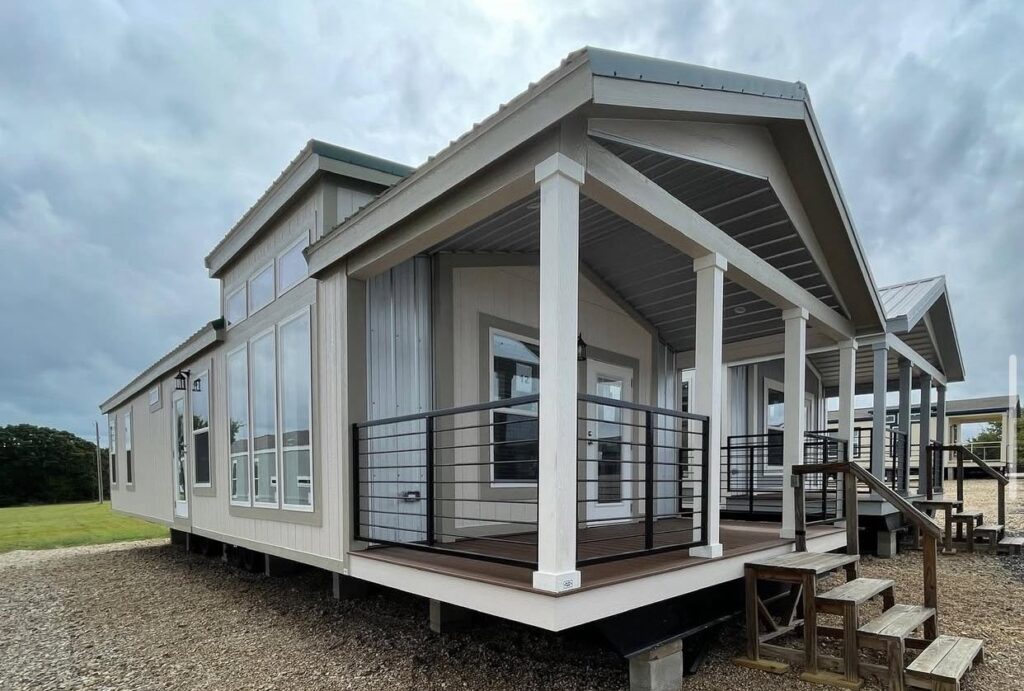 Modern tiny house with large windows, grey siding, and a covered porch. The overcast sky and surrounding greenery create a cozy, serene ambiance.