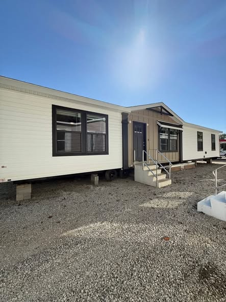 A mobile home on a gravel lot under a clear blue sky. It has a modern design with white siding, large black-framed windows, and a small front porch.