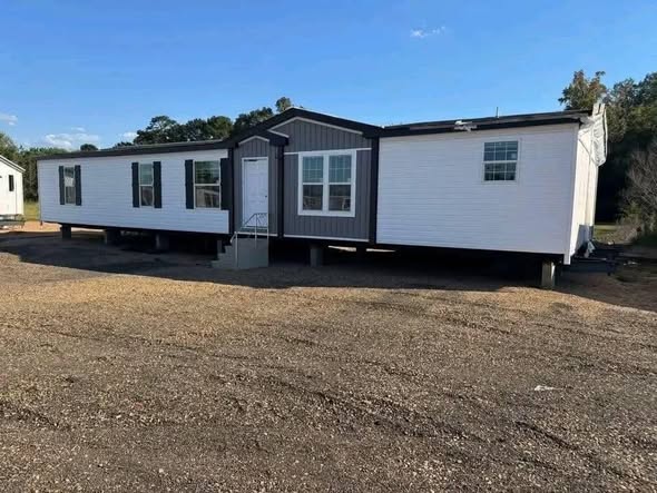 A white modular home with a dark trim sits elevated on a gravel lot, surrounded by trees under a clear blue sky, conveying a calm, rural setting.