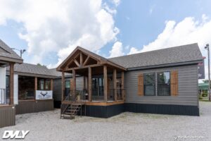 Modern gray tiny house with wooden accents and gable porch under a partly cloudy sky. Gravel ground adds a rustic touch. Cozy and inviting atmosphere.
