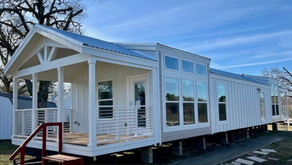 A modern, white modular home with large windows and a metal roof sits on a sunny day. The porch has a railing and is accessed by red steps.