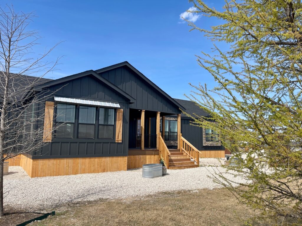 Black modern farmhouse with wooden accents, elevated porch, and gravel pathway. Surrounded by sparse trees under a clear, blue sky. Calm and inviting.