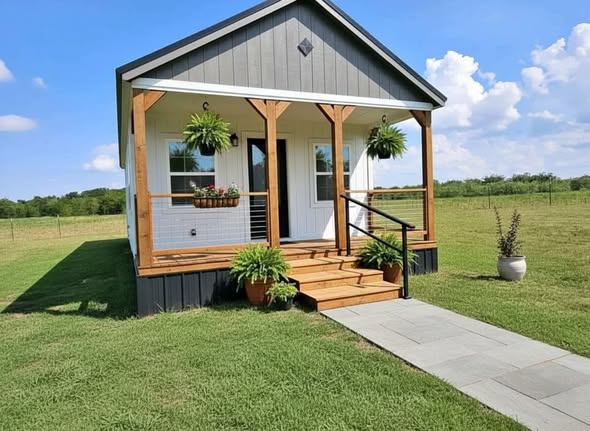 A quaint tiny house with a gray exterior and welcoming porch adorned with hanging plants. Set in a lush green field under a bright blue sky.