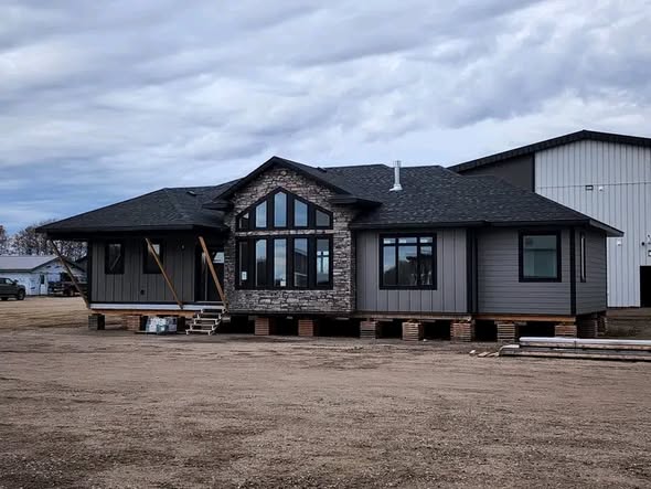 Single-story house under construction sits on blocks, featuring gray siding, large windows, and a stone facade. Overcast sky creates a somber tone.