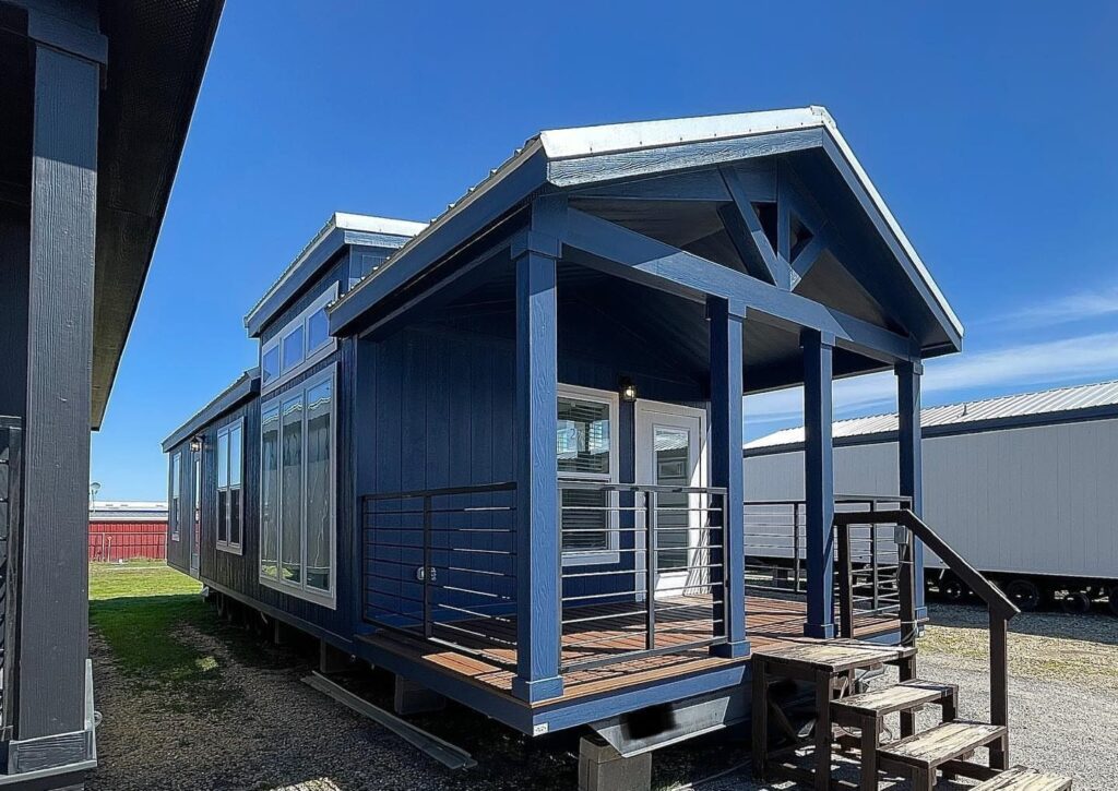 A modern tiny house on display features a dark blue exterior and large windows. It has a covered porch with metal railings and steps leading up. The scene conveys a sense of sleekness and innovation under a clear blue sky.