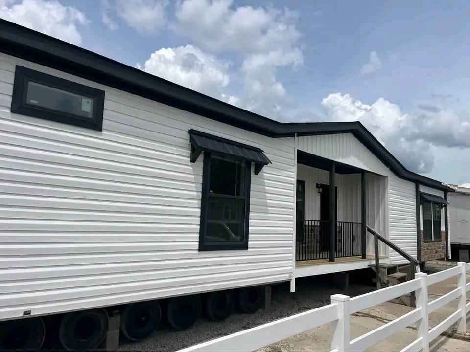A modern manufactured home with white siding and black trim under a partly cloudy sky. Features include a small porch with a railing and steps.