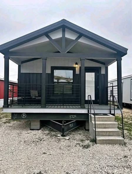 A modern tiny house with a gabled roof and dark trim is elevated on a platform. It features a small porch with railings and steps leading down.